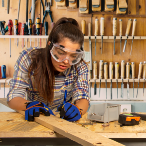 Una mujer que lleva gafas de seguridad ventiladas antivaho mientras trabaja con madera y herramientas para madera en su estudio de arte