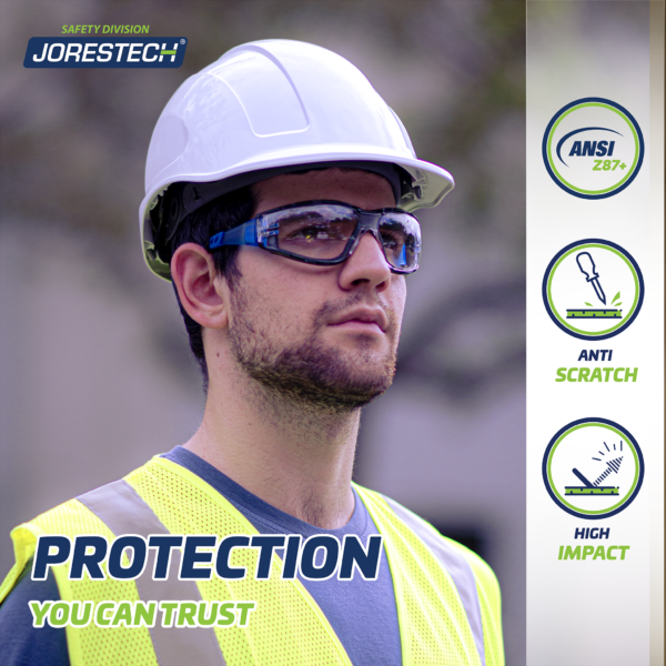 Un trabajador con chaleco reflectante color lima, casco de seguridad y las gafas de seguridad transparentes JORESTECH antiempañantes para protección de alto impacto. El texto dice: Protección en la que puede confiar. Los iconos indican: antiarañazos, alto impacto, cumple con ANSI Z87+