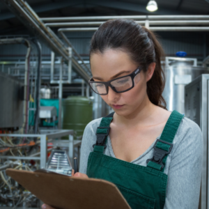Una mujer trabajando en una fábrica. Lleva puestas las gafas de lectura de seguridad bifocales JORESTECH con protecciones laterales para protección contra impactos fuertes mientras toma notas en un portapapeles.