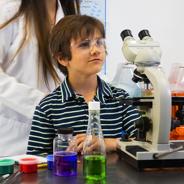 Un niño que lleva gafas de seguridad transparentes JORESTECH para niños, para protección contra alto impacto, en un entorno de laboratorio. Está usando un microscopio y tiene una gran variedad de frascos con líquidos de colores vivos para examinar. Su maestra está al fondo.