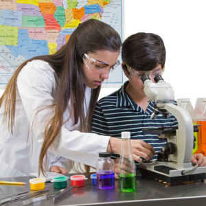 Una profesora en su laboratorio usando las gafas superpuestas transparentes Jorestech, conformes con ANSI, mientras ayuda a un niño con un microscopio. Mostradas en un entorno de aula.