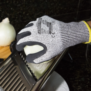 Manos de una persona que lleva los guantes de trabajo de seguridad negros JORESTECH resistentes a cortes con palma recubierta de nitrilo mientras corta una cebolla con un cortador de cuchillas de metal plateado