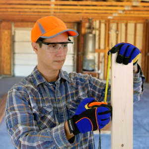 Hombre en un garaje con guantes de seguridad y las gafas de seguridad rectangulares con montura y protectores laterales para protección ocular contra impactos altos, midiendo un trozo de madera que usa en un proyecto de bricolaje