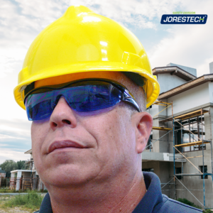 Un hombre de pie fuera de una construcción de casas adosadas. Lleva gafas de seguridad envolventes de alto impacto JORESTECH azules y un sombrero amarillo estilo gorra. Hay varios andamios colocados frente a las casas para instalar ventanas y continuar pintando.