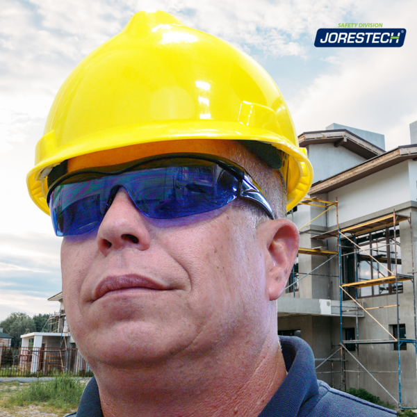 Un hombre de pie fuera de una construcción de casas adosadas. Lleva gafas de seguridad envolventes de alto impacto JORESTECH azules y un sombrero amarillo estilo gorra. Hay varios andamios colocados frente a las casas para instalar ventanas y continuar pintando.