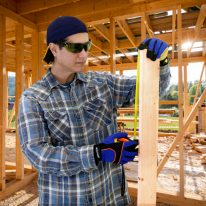 Hombre usando las gafas de seguridad envolventes con montura verde mientras mide una tabla de 2 x 4 para continuar con la construcción de una casa. La casa aún no tiene paredes, por lo que el trabajador usa estas gafas tintadas para protegerse del sol.