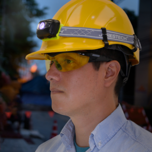 Un trabajador que lleva un casco amarillo con una lámpara frontal sujeta a él con los clips para lámpara frontal de casco Jorestech. El fondo es un desenfoque oscuro con señales de tráfico.