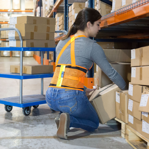 Una mujer lleva un cinturón de soporte para la espalda ajustable de alta visibilidad con bandas reflectantes mientras carga una caja en un almacén. Se aprecia el color naranja brillante y las correas ajustables en la parte trasera.