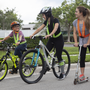 Muestra a un niño y 2 jóvenes andando en bicicleta y scooter en la carretera usando los tirantes de seguridad de alta visibilidad para jugar