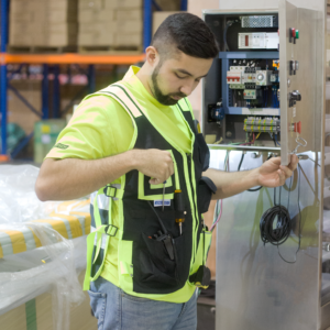 Un electricista que lleva el chaleco portaherramientas lleno de diferentes herramientas. Está trabajando en el panel de circuitos de una máquina