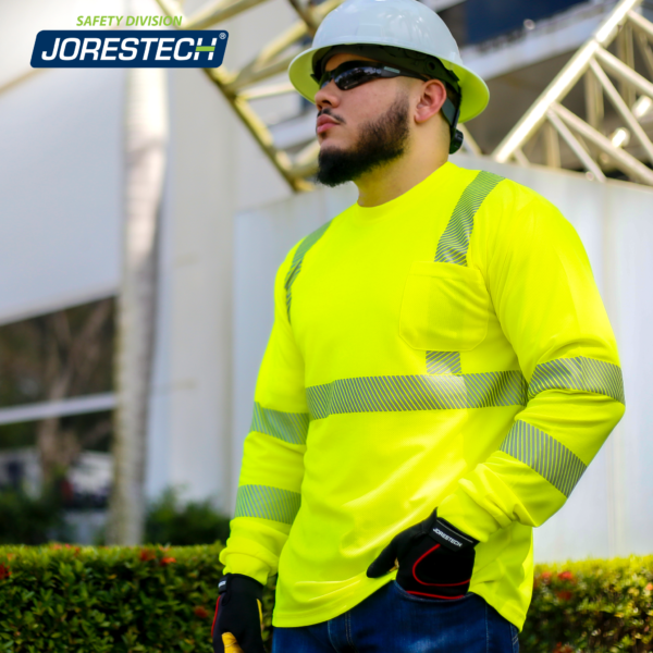 Un trabajador de pie cerca de una estructura de metal mientras lleva la camiseta de manga larga de seguridad JORESTECH amarilla de alta visibilidad con transferencia térmica reflectante, un casco blanco y guantes de trabajo negros