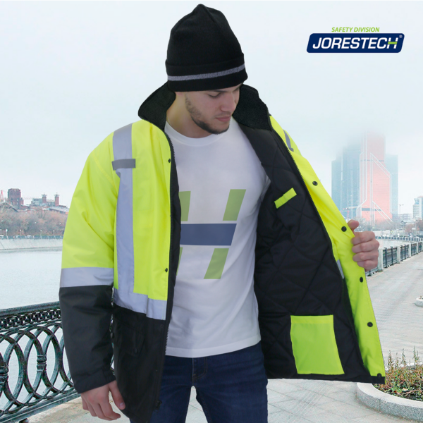 Hombre en una ciudad que lleva la chaqueta parka de seguridad JORESTECH de alta visibilidad en amarillo y negro, conforme a ANSI, con bolsillos y franjas reflectantes. El hombre está buscando algo dentro de los bolsillos ubicados en la capa interior de la chaqueta parka de seguridad.