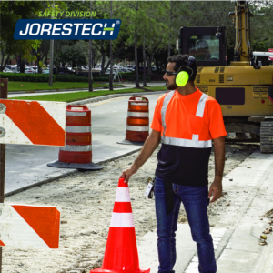 Un trabajador de pie en medio de una obra vial mientras lleva la camisa de seguridad naranja reflectante JORESTECH. Hay un gran camión amarillo al fondo