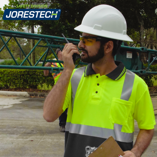 Trabajador hablando por un walkie-talkie y vistiendo la polo reflectante de alta visibilidad. Gran caja en el fondo y otros trabajadores.