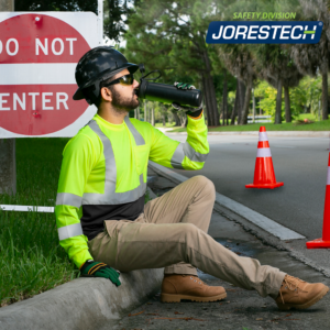 Un trabajador que lleva una camisa de seguridad JORESTECH de alta visibilidad y un casco de seguridad negro mientras bebe agua al borde de la carretera