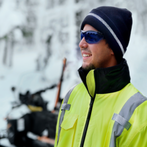 Un trabajador que lleva el gorro beanie reflectante negro JORESTECH® y una chaqueta de alta visibilidad mientras conduce una moto de nieve