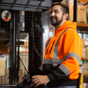 Trabajador que lleva una sudadera de seguridad JORESTECH naranja con franjas reflectantes mientras conduce una carretilla elevadora
