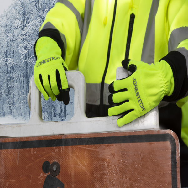 Persona usando los guantes de seguridad hi-vis JORESTECH para mover una señal de precaución de calle de un lugar a otro en un entorno invernal para construcción vial