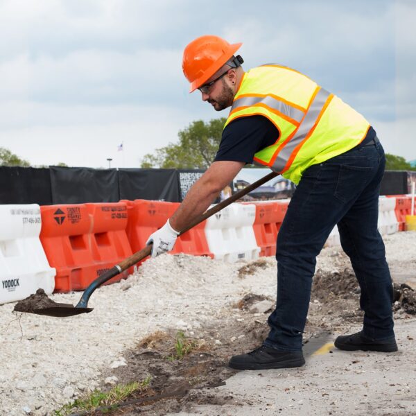 Imagen de un hombre con un chaleco de seguridad JORESTECH bicolor en naranja lima, guantes de trabajo y casco de seguridad naranja, trabajando en una obra cerca de la carretera