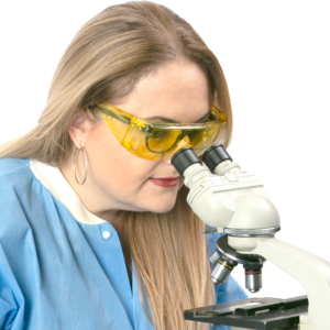 Una mujer con EPP en un entorno de laboratorio llevando las gafas de seguridad Jorestech amarillas sobre sus gafas graduadas. La mujer está mirando a través de un microscopio