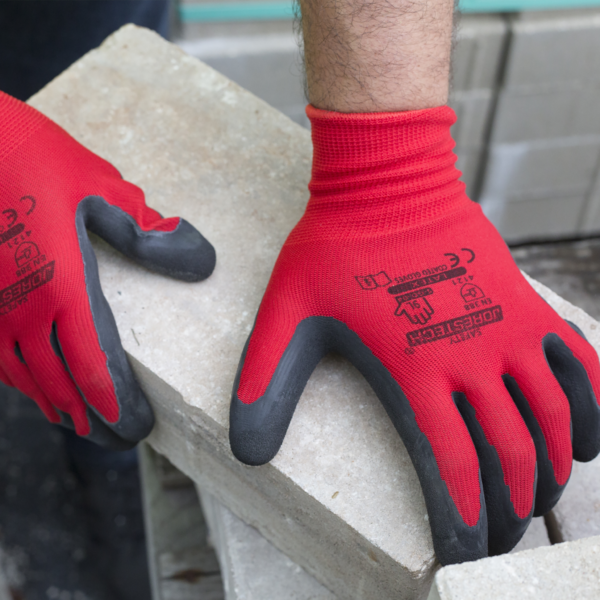 Manos de un trabajador que lleva los guantes de trabajo JORESTECH rojos y negros con palmas recubiertas de látex, usados en construcción para transportar pesados ladrillos rústicos de cemento