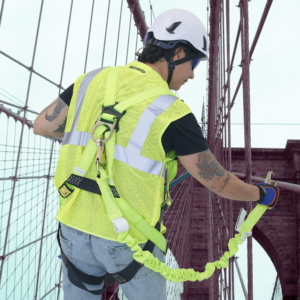 Un trabajador con casco blanco con barboquejo y un arnés con una sola pata de cabo con absorbedor interno de energía, enganchado al anillo en D del arnés. Se está asegurando a una parte estable de la estructura de un puente durante tareas de mantenimiento o reparación a gran altitud.