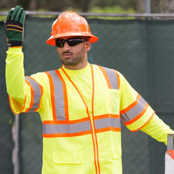 Un trabajador dirigiendo el tráfico con ropa reflectante de alta visibilidad amarilla, un casco de seguridad naranja y gafas de seguridad JORESTECH Smoke para protección de alto impacto durante una construcción vial.